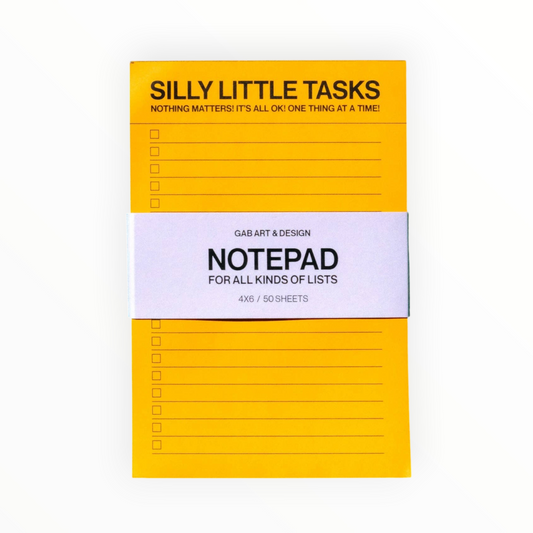 A yellow notepad with black printed text reading 'SILLY LITTLE TASKS' and 'NOTEPAD FOR ALL KINDS OF LISTS' on a white paper band, photographed on a white background.
