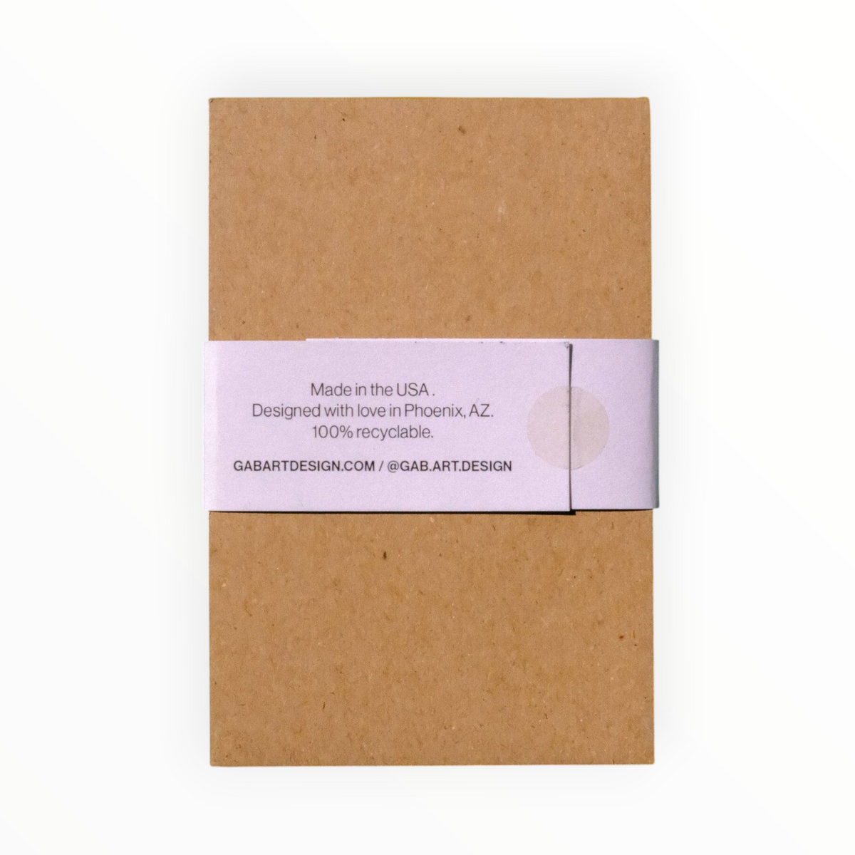 A square kraft paper notebook or journal wrapped with a light lavender paper band that reads 'Made in the USA. Designed with love in Phoenix, A.