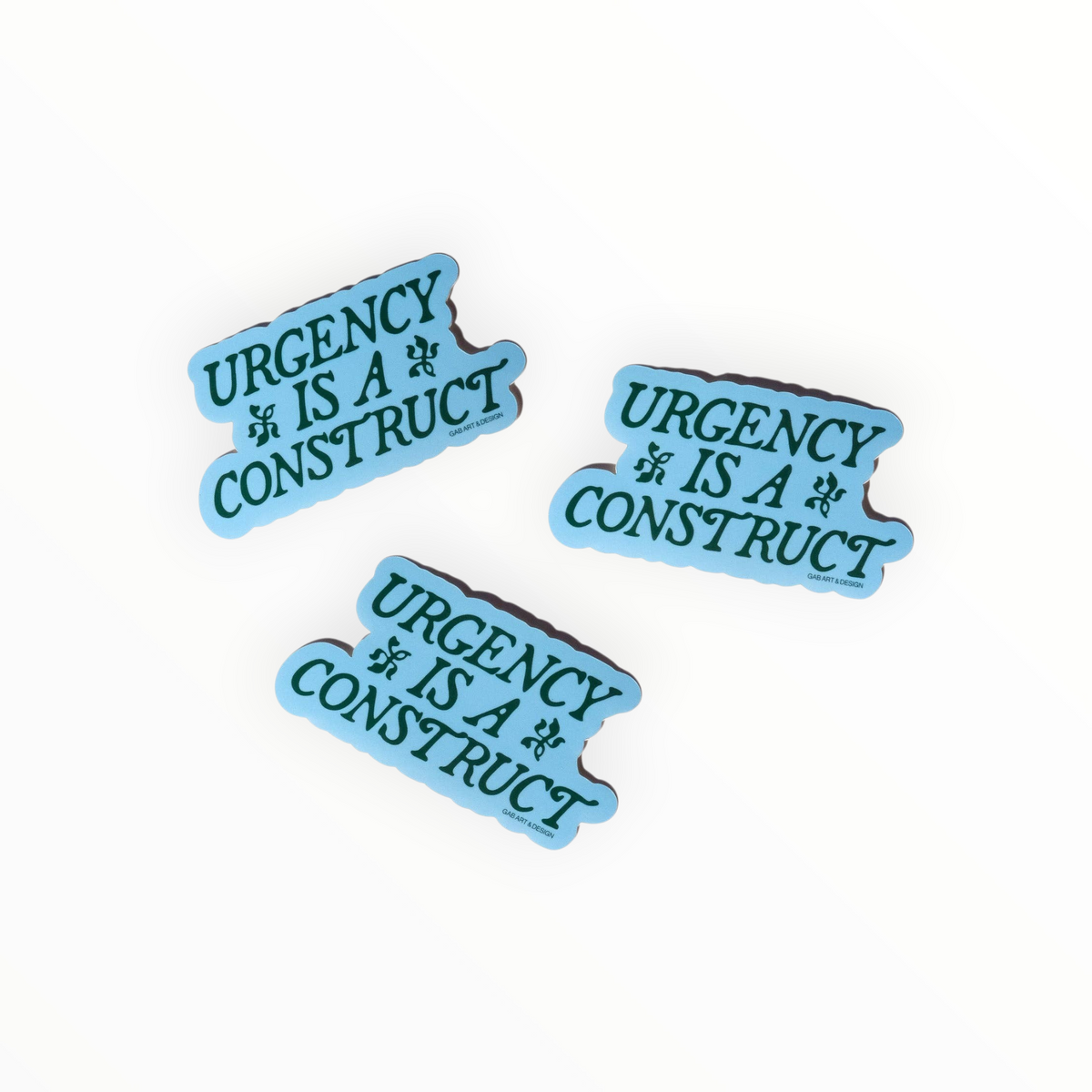 Three matching light blue enamel pins with the text 'URGENCY IS A CONSTRUCT' in dark teal lettering, arranged on a plain white background.